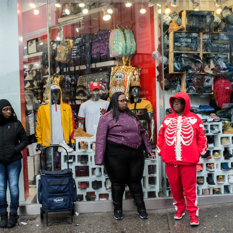 March 30, 2020: One person stands wearing drab winter clothes in contrast with the studied flamboyance of the other two. One wearing a jogging outfit with a rib cage design. Notice the use of cinderblocks for displaying sneakers. Dr. Jay’s, 410 Westchester Avenue, Bronx, New York. © Camilo José Vergara 